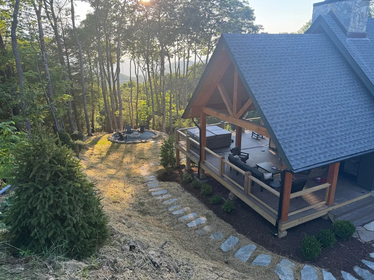 A charming wooden cabin structure with an inviting outdoor seating area surrounds a classic stone fire pit. A stone pathway leads through lush trees, highlighting spaces for storytelling, stargazing, and outdoor feasts.