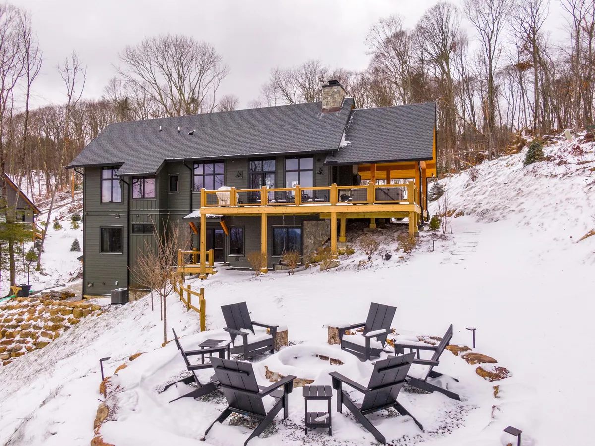 A large wooden deck with a modern house backdrop, featuring a welcoming fire pit area with seating. The deck is set against a wintery landscape, suggesting year-round use and cozy gatherings by the fire.