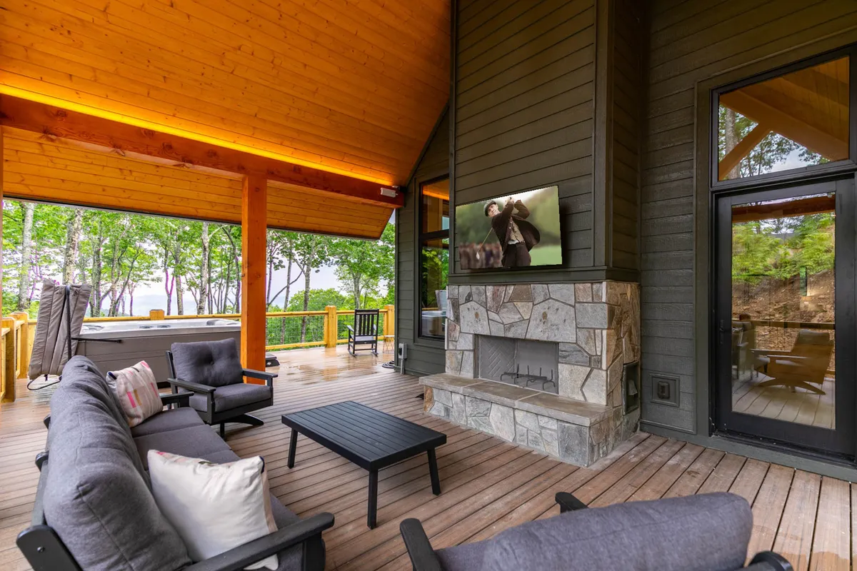 An inviting outdoor seating area with a stone fireplace, televisions, and a hot tub, all set under wooden beams. The area is clearly designed for entertaining, with both relaxation and social activities in mind.