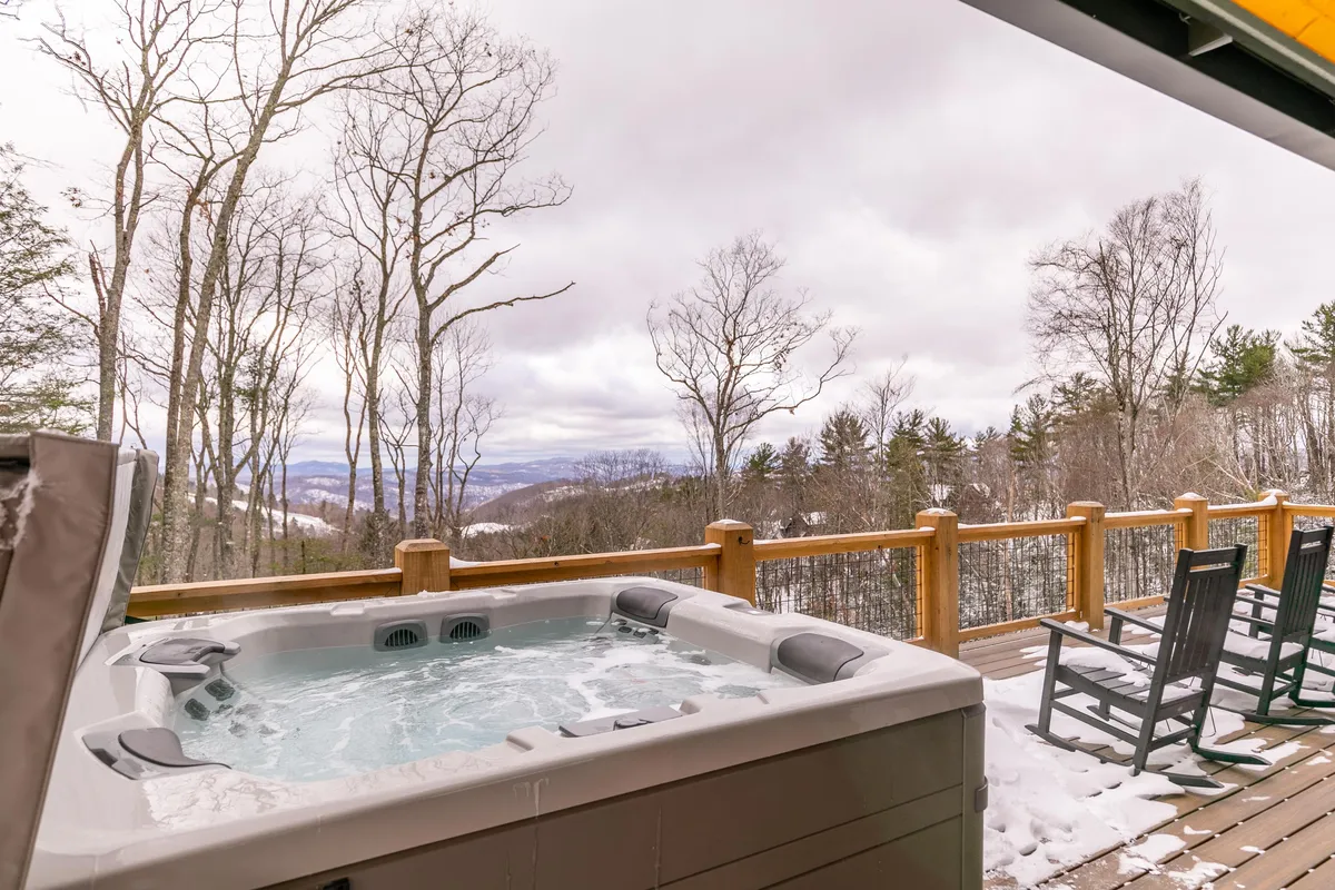 An inviting deck with a hot tub, wooden railing, and rocking chairs overlooks distant mountains and bare trees, suggesting a cozy and serene retreat in cooler months.