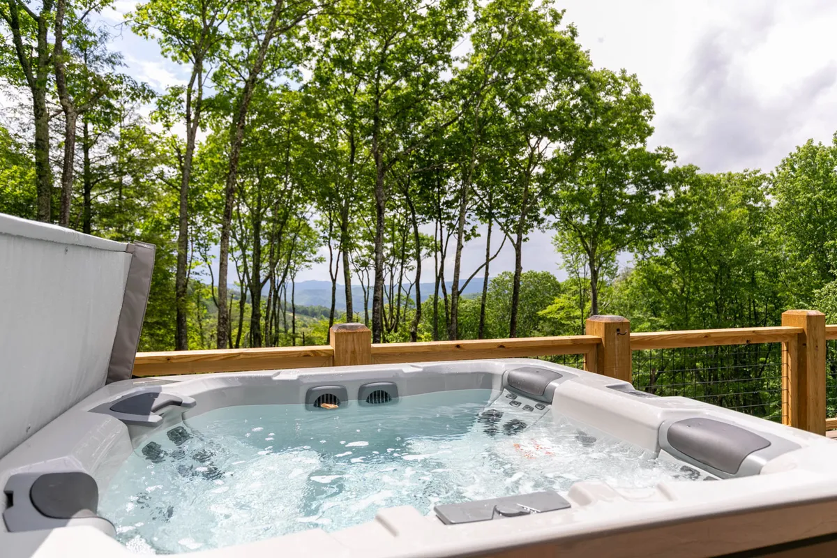 A serene view of a private outdoor hot tub nestled among tall trees, with distant mountain vistas. The hot tub area is surrounded by nature, and the setting exudes tranquility and luxury—perfect for relaxing after a day of exploration.