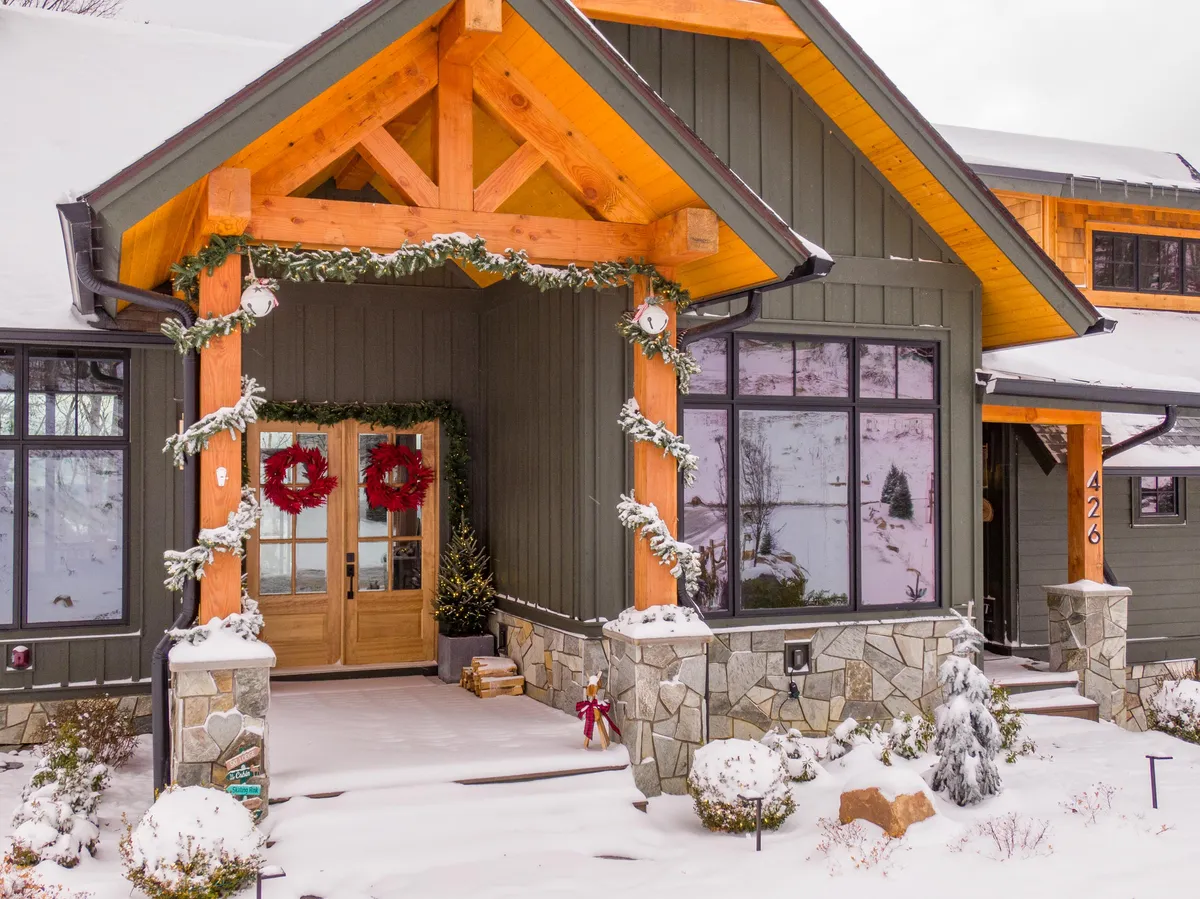 A snow-covered cabin entry framed by wooden double doors adorned with wreaths, garland, and festive ornaments. Stonework at the base and a soft layer of snow set a holiday atmosphere, while the decorative elements add to the wintertime ambiance.