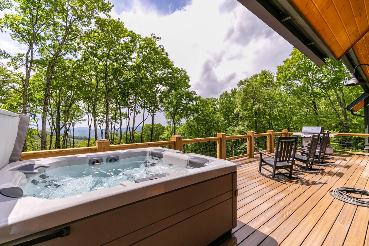 A tranquil outdoor deck featuring a bubbling hot tub, wooden deck, rocking chairs, and expansive wooded and mountain views. The scene invites relaxation and is set up for both entertainment and quiet enjoyment, with the hot tub as a central focal point.