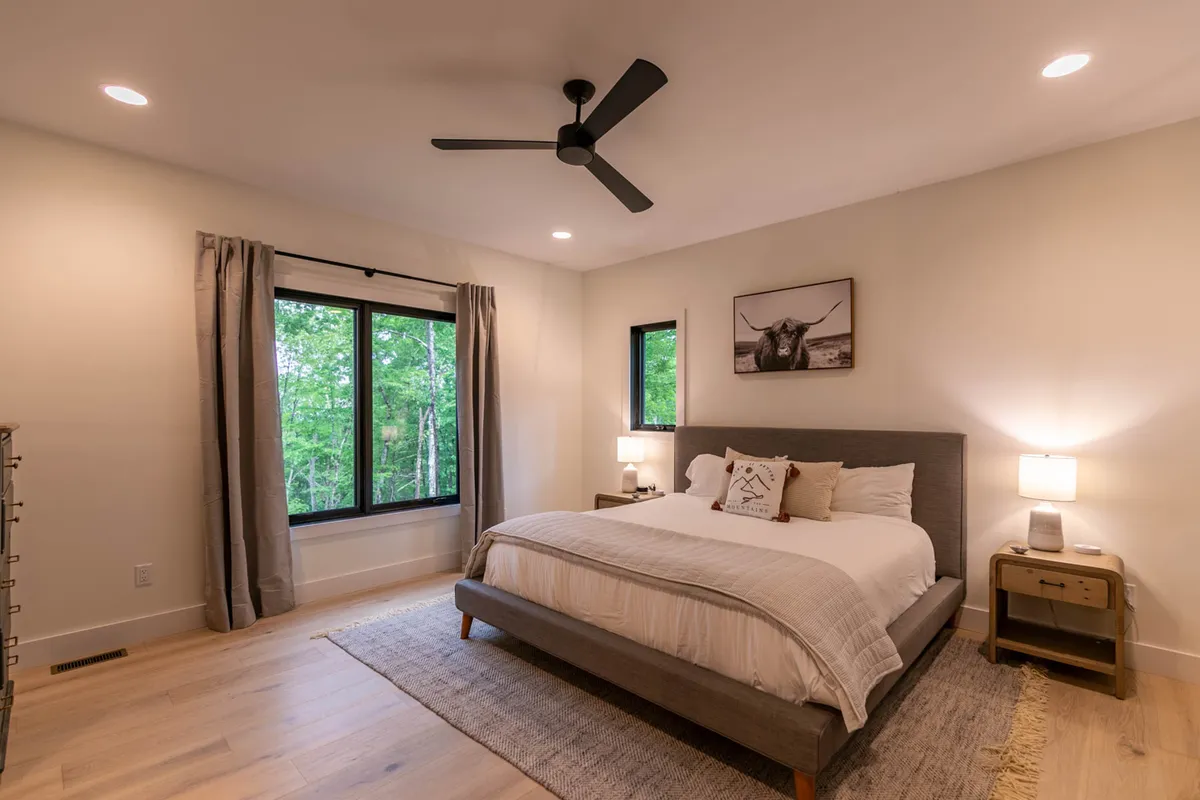 A serene bedroom with a plush king bed adorned with decorative pillows, flanked by large windows offering tranquil views of the surrounding nature. The room features artwork on the wall and cozy reading nooks, creating an inviting and calming retreat.