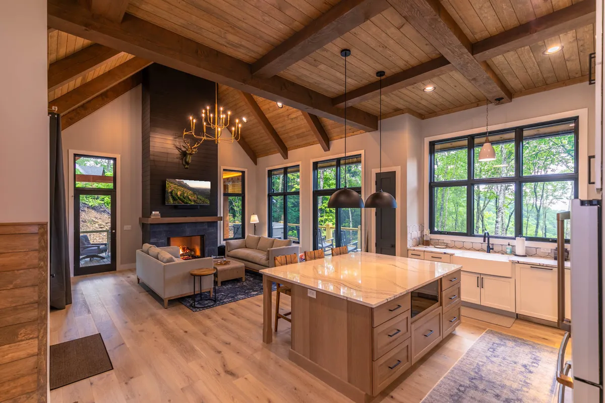 An open-concept living area featuring a spacious kitchen with a large central island, plush sofa seating, a cozy fireplace, and expansive windows offering views of the surrounding nature. The space is illuminated by natural light and designed for social gatherings and family relaxation.