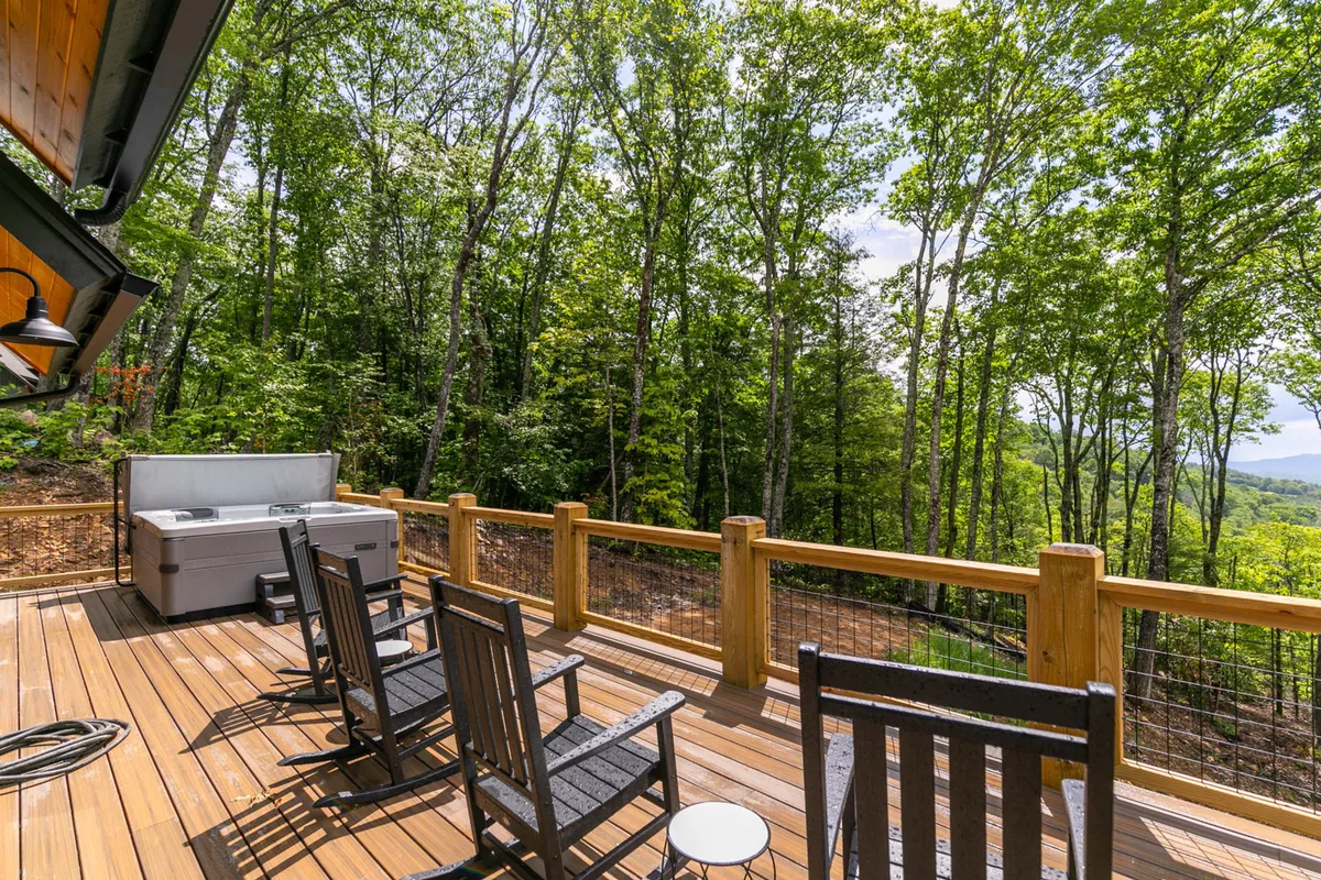 A serene outdoor deck featuring a wooden hot tub, surrounded by tall trees. Rocking chairs are arranged on the deck, offering inviting spots to relax after a day outdoors.