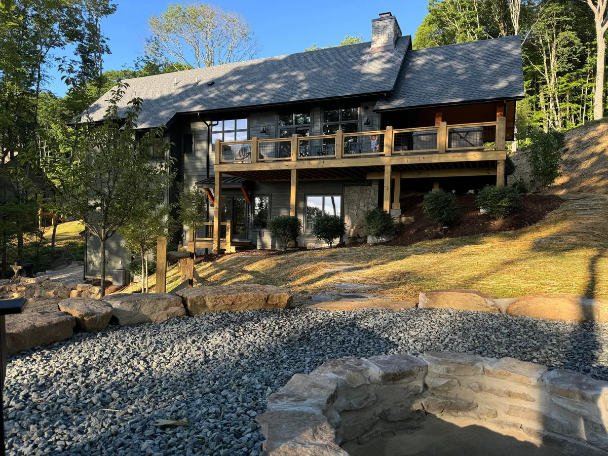 A tranquil deck with wooden railings overlooks a manicured garden and features a prominent stone fire pit, inviting outdoor dining and relaxation. Large windows connect the deck to the home, while comfortable seating around the fire pit sets the stage for evening gatherings under the stars.