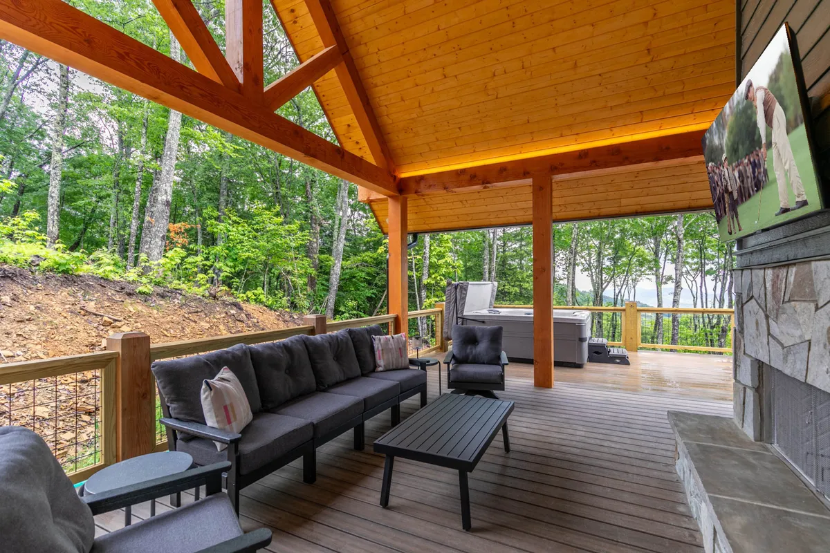 A cozy outdoor seating area featuring a hot tub and a television mounted on a stone wall is set beneath a protective wooden ceiling. The area is surrounded by serene nature views, making it ideal for both lively group activities and tranquil relaxation.